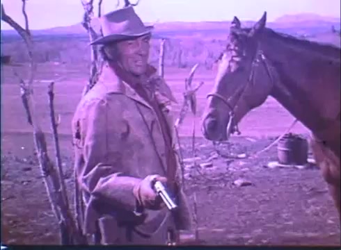 The still from the 'Showdown' trailer features a rugged cowboy, likely portraying a central character, holding a revolver with a confident expression. He wears a wide-brimmed hat and a weathered coat, standing next to a horse in a barren landscape. The colors reflect the vintage quality of the 16mm print, enhancing the Western ambiance. The scene conveys tension and anticipation typical of Western films.