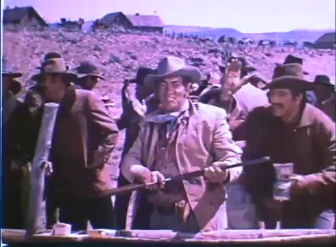 The still from the 'Showdown' trailer features a tense scene with a group of men in cowboy attire, standing outdoors, likely in a Western setting. The central figure, holding a rifle, appears ready for a confrontation, while others look on, some with drinks in hand. The landscape is rugged, with wooden structures in the background, hinting at a small-town atmosphere typical of Western films. The overall mood suggests a classic standoff moment.