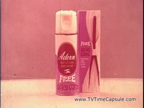 The still from the 16mm print features a can of Adorn Red Styling Hair Spray prominently displayed alongside a package promoting a 'Free Kicky Comb.' The colors are soft and muted, typical of the era, with a nostalgic feel. The packaging emphasizes the product's benefits, suggesting it provides lasting hold. The overall presentation reflects 1970s advertising aesthetics, focusing on self-styling and hair care.