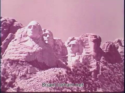 The still features a view of Mount Rushmore, showcasing the carved faces of four U.S. presidents: George Washington, Thomas Jefferson, Theodore Roosevelt, and Abraham Lincoln. The image is styled in a vintage color palette, typical of 1960s film, highlighting the monumental nature of this national landmark. The context suggests a celebration of American heritage and iconic landmarks in a public service announcement aimed at promoting tourism.
