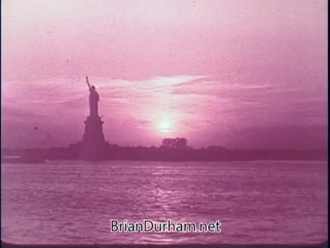 The still from the 'Discover America PSA - 1969' features the Statue of Liberty silhouetted against a vibrant sunset. The scene is dominated by soft pink and purple hues in the sky, reflecting on the water's surface. This serene composition conveys a sense of national pride and beauty associated with American landmarks.