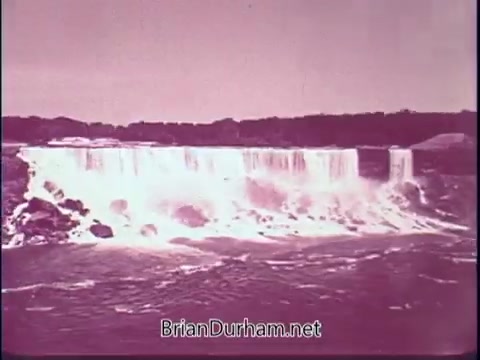 The still features a serene view of Niagara Falls, showcasing the powerful cascade of water. The image has a vintage quality with a pinkish hue, characteristic of 16mm film from the late 1960s. The scene emphasizes the natural beauty and majesty of the falls, fitting the promotional theme of exploring American landscapes in the 'Discover America' campaign.