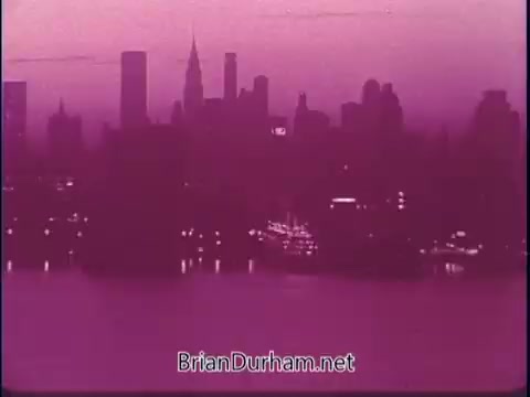 The still features a silhouetted city skyline at dusk, showcasing iconic buildings like the Empire State Building. The scene is bathed in a purplish hue, contributing to a nostalgic and atmospheric feel. It represents a moment of calm over a bustling urban landscape, likely aiming to evoke a sense of wonder and appreciation for America’s cities in the context of the PSA.