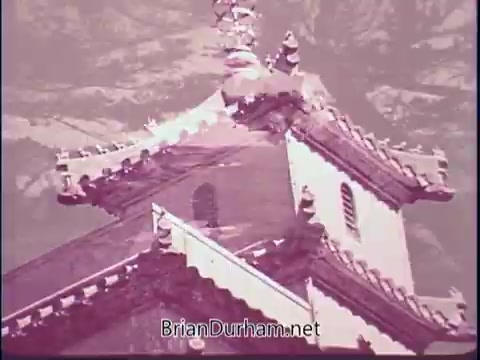 The still from the 'Discover America PSA - 1969' features a close-up view of an ornate building, likely showcasing architectural details including intricate rooflines and decorative elements. The color palette appears muted, adding a vintage feel to the image. The backdrop suggests a natural setting, possibly mountainous, enhancing the sense of exploration and scenic beauty associated with the campaign.