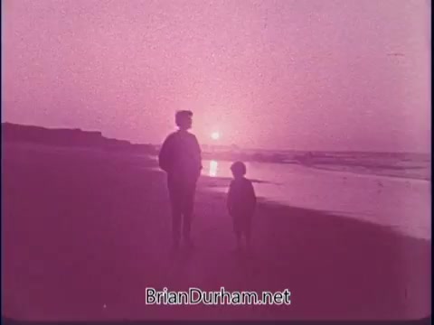 The still from the 'Discover America' PSA features a silhouetted adult and child standing on a beach, gazing at a sunset. The scene is bathed in a pink hue, creating a nostalgic and emotional atmosphere. The ocean waves can be seen softly lapping at the shore, emphasizing a tranquil and reflective moment.