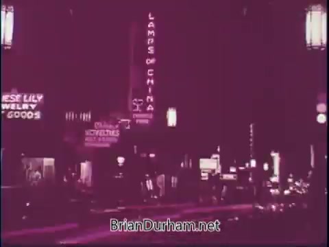 The still from the 'Discover America PSA - 1969' showcases a vibrant urban street scene at night. Neon signs, such as 'LAMPS OF CHINA' and other storefronts, illuminate the surroundings, creating a lively atmosphere. The color palette conveys a nostalgic feel, emphasizing the energy and character of American city life during that era.