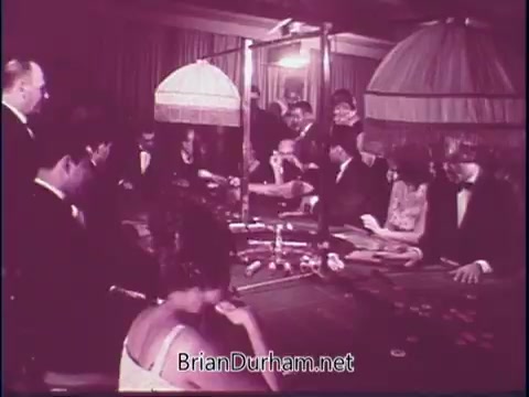 The still depicts a dimly lit casino scene featuring a group of elegantly dressed individuals gathered around a gaming table. Men in tuxedos and women in formal attire engage in a game, with chips and cards present. The atmosphere is lively and sophisticated, characterized by vintage elements typical of the late 1960s. The presence of unique table lamps adds to the ambience.