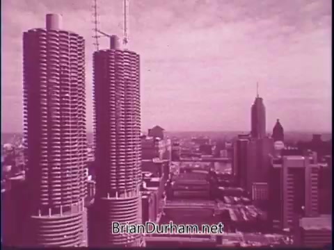 The still features a panoramic view of a city skyline, prominently showcasing two cylindrical towers with distinctive textures, likely representing modern architecture. The scene conveys a nostalgic aesthetic, typical of 1960s film, with a pinkish tint. In the background, there are additional skyscrapers, including a recognizable tall structure suggesting urban vibrancy. This image likely captures the essence of American city life during that era, emphasizing architectural innovation and urban development.