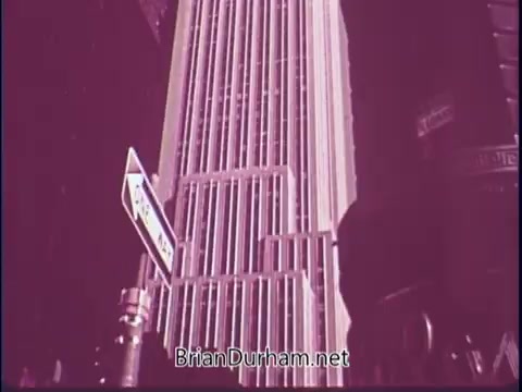 The still from 'Discover America PSA - 1969' features a tall, modern skyscraper with prominent vertical lines. A street sign is visible in the foreground, suggesting an urban setting. The overall tone of the image is a washed-out pink, giving it a vintage aesthetic characteristic of 1960s film. The scene invokes a sense of exploration and appreciation for American architecture.