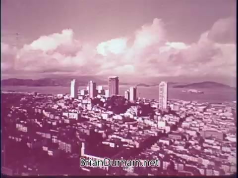 The still from the 1969 'Discover America PSA' showcases a panoramic view of a cityscape, likely depicting a modern urban area with tall buildings. The image features a blend of cloudy skies and water in the background, suggesting a coastal location. The color palette is muted, contributing to a nostalgic or vintage feel typical of the era. The scene effectively captures the essence of American urban life and invites viewers to explore the beauty of the country.