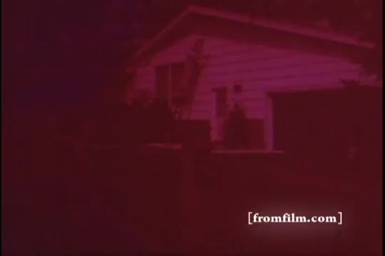 The still from the 'Ajax White Tornado' commercial features a house, predominantly colored in deep red hues typical of a 16mm print’s color palette. The structure appears simple and suburban, with some landscaping visible. This still likely captures a moment emphasizing cleanliness or a relatable domestic scene, aligning with the commercial's theme of household products. The overall aesthetic conveys a vintage feel, characteristic of mid-20th century advertising.