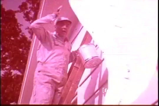 The still features a man in a light-colored jumpsuit and cap, standing on a ladder while holding a paint bucket. He appears to be engaged in a painting task on the side of a house. The image has a vintage quality typical of a 16mm film, capturing a moment of home improvement, likely promoting the cleaning power of Ajax White Tornado. The lighting gives it a warm, nostalgic tone.