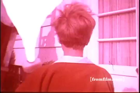 The still from the 'Ajax White Tornado' commercial features a child with tousled hair, seen from the back. The scene is tinged in a pink hue, capturing a playful and bright atmosphere. In the foreground, a large white object—likely representing the Ajax product—dominates the image, contrasting against the backdrop of a house with a window. This visual creates a sense of excitement and approachability, typical of mid-20th century advertising aesthetic.