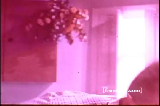 The still from the Ajax White Tornado commercial features a softly lit interior scene with a focus on a quilted bed and a wooden shelf adorned with flowers. The color palette is warm, with a reddish tint, suggesting a cozy home atmosphere. The composition hints at a domestic setting, likely as part of a narrative promoting Ajax cleaning products.