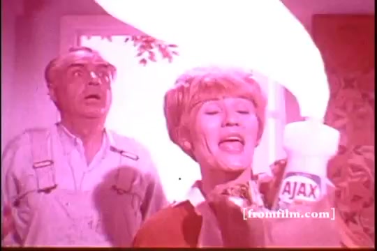 The still features a woman enthusiastically promoting Ajax cleaning product, holding a bottle with excitement. She has a lively expression, gesturing as if demonstrating its effectiveness. In the background, a man appears surprised or shocked, enhancing the dramatic moment. The overall scene is bright and colorful, typical of vintage commercials, likely conveying the product's transformative power in a humorous way. The color palette has a warm, pinkish hue, characteristic of the 16mm film format.