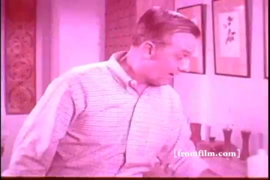 The still features a man in a light-colored, collared shirt, appearing surprised or intrigued as he interacts with items on a surface. The background suggests a domestic setting, with decorative elements like artwork on the walls. The color palette has a pinkish hue, typical of older 16mm film prints, adding a vintage feel to the scene. The context is from a commercial promoting Ajax White Tornado, likely focusing on its cleaning power.
