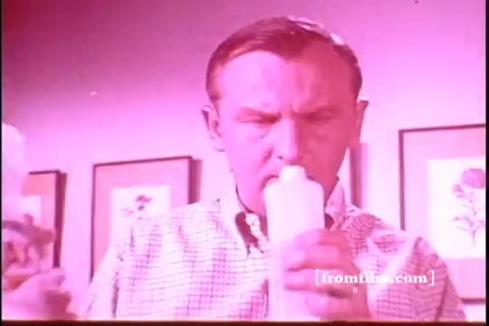 The still from the Ajax White Tornado commercial features a man intently examining a bottle of Ajax cleaner. He appears to be in a domestic setting, characterized by framed floral artwork on the walls. The lighting has a pinkish hue, typical of certain vintage film prints. The man's expression suggests curiosity or concentration as he engages with the product.