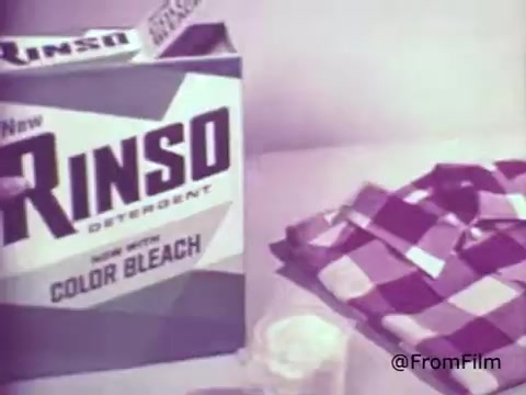 This still from a 1960s commercial for Rinso Detergent showcases a box prominently featuring the product name and the phrase 'Now with Color Bleach!' The box is designed with bold colors typical of that era. Nearby, a checkered fabric is displayed, emphasizing the detergent's effectiveness on colored laundry. The overall composition reflects the advertising style of the time, aiming to attract consumers seeking vibrant cleaning solutions.   For more vintage film insights, follow us on Twitter @FromFilm.