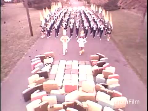 The still from the 1979 Samsonite Luggage commercial features a lively scene with a marching band and colorful flags in formation on a road. In the foreground, various pieces of Samsonite luggage are arranged in a block pattern, leading to the marching group. The setting appears outdoors, possibly in a park-like area, suggesting a festive or promotional atmosphere. The color palette is soft and vintage, typical of the era's film techniques.