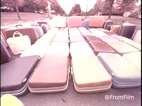 The still from the 1979 Samsonite luggage commercial displays a vibrant array of suitcases arranged neatly on a street. The suitcases, in various colors such as brown, tan, and pastel shades, are stacked in multiple rows, showcasing the variety of styles offered by Samsonite. The scene is set against a backdrop of suburban trees, conveying a sense of travel and adventure. The vintage aesthetic reflects the era's marketing style, emphasizing functionality and durability.