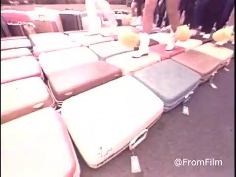 The still from the 1979 Samsonite luggage commercial shows a colorful array of luggage pieces arranged on the ground, possibly as part of a promotional event. Several individuals stand on or near the suitcases, with some holding pom-poms, suggesting an upbeat atmosphere. The visual emphasizes the variety of Samsonite luggage, showcasing its design and appeal. The shot captures a lively scene, likely aimed at drawing attention to the brand.