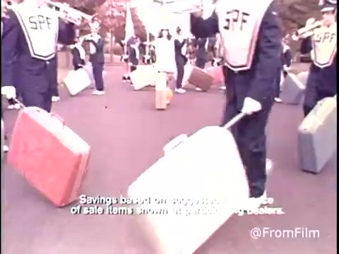 The still from the 1979 Samsonite Luggage commercial features a group of individuals, likely performers, in uniforms that prominently display 'SPF.' They are shown carrying various pieces of luggage, including hard-shell suitcases in different colors. The setting appears to be outdoors, possibly during a promotional event or parade. Below the image, there's text mentioning savings based on specific items shown, indicating a focus on marketing the luggage. The overall vibe is energetic and promotional, typical of commercials from that era.