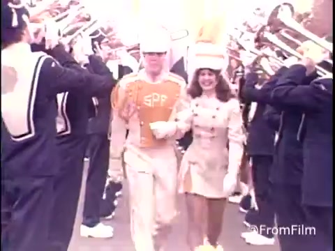 The still from the 1979 Samsonite Luggage commercial features a parade atmosphere. In the foreground, a marching band member in an orange and white uniform walks alongside a female performer in a light-colored outfit. Both appear cheerful and confident as they make their way through a lineup of musicians playing brass instruments. The background is filled with band members, creating a lively and energetic scene. The overall tone suggests celebration and excitement, aligning with the vibrant spirit often portrayed in commercials of that era.
