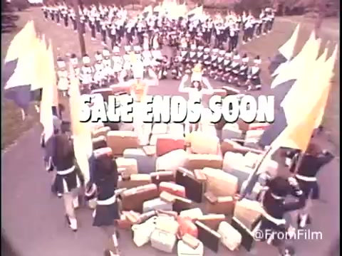The still from the 1979 Samsonite luggage commercial features a vibrant promotional scene. In the foreground, an array of colorful luggage is arranged in a circular pattern. Surrounding the luggage, a group of performers dressed in uniforms and holding flags contributes to the festive atmosphere. The text 'SALE ENDS SOON' prominently displays the urgency of the promotion, capturing the viewer's attention against the backdrop of the performance. The overall composition conveys excitement and encourages potential customers to take advantage of the sale.