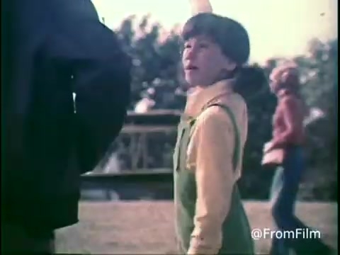 The 16mm still from the 1970s Kellogg's Shredded Wheat commercial features a young child in a green outfit with a yellow long-sleeve underneath, looking up attentively at an adult figure. In the background, another child can be seen wearing a red jacket, hinting at a playful outdoor setting. The image captures a moment of engagement, emphasizing the wholesome, family-friendly appeal of the brand. The natural lighting and casual attire evoke a nostalgic vibe typical of the era.   Follow us on Twitter @FromFilm.
