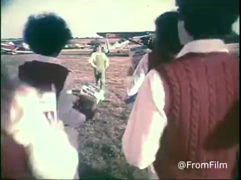 In this still from the 1970s Kellogg's Shredded Wheat commercial, a group of children, wearing red vests, enthusiastically approaches a figure dressed in bright yellow. The backdrop features vintage airplanes and an open field, capturing a playful and nostalgic vibe typical of the era. The scene likely evokes themes of adventure and fun associated with the cereal brand.   Follow us on Twitter @FromFilm.