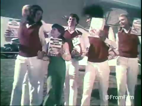 The still features a group of cheerful kids and young adults dressed in matching outfits with red vests and white pants, joyfully holding up boxes of Kellogg's Shredded Wheat. They appear to be in a sunny outdoor setting, likely promoting the cereal in a fun, energetic manner. The vibrant colors and playful expressions reflect the lively spirit typical of 1970s commercials.