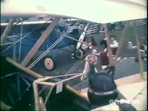The 16mm still from the 1970s Kellogg's Shredded Wheat commercial features a scene set by vintage biplanes. In the foreground, individuals are engaged with the aircraft, likely depicting a playful or adventurous theme. The overall color palette has a nostalgic feel typical of that era, enhancing the lively atmosphere. The image captures a moment of engagement, hinting at a fun, family-oriented concept promoting the cereal.