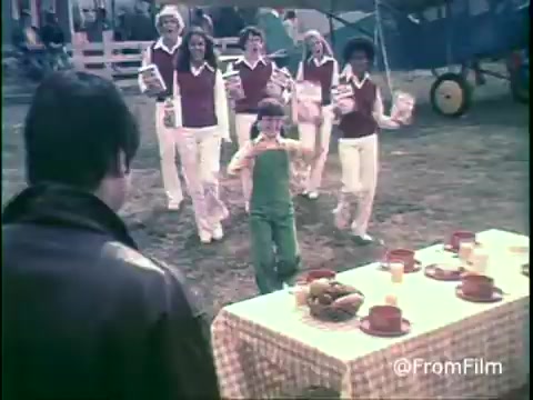 The still features a scene from a 1970s Kellogg's Shredded Wheat commercial. A child in green overalls joyfully runs toward a table set with breakfast items, while a group of adults in matching outfits with red vests cheerfully approaches from behind. The setting appears to be an outdoor event, with a picnic-style atmosphere and colorful props. The print's vintage aesthetic reflects the era's advertising style.