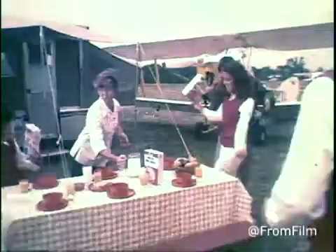 The still captures a vibrant outdoor scene featuring a picnic setup with a checkered tablecloth. Two women are engaged with food preparation near a table adorned with bowls of Kellogg's Shredded Wheat and other breakfast items. A tent and some campers are visible in the background, suggesting a casual, communal atmosphere typical of 1970s family gatherings. The color palette has a nostalgic, faded quality characteristic of 16mm film prints. The overall vibe is playful and cheerful, aligned with the era's advertising style.