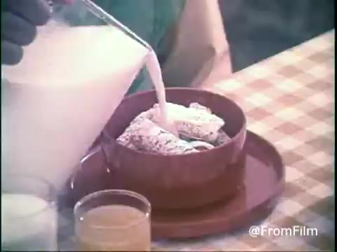 The still features a hand pouring milk into a bowl of Kellogg's Shredded Wheat, set on a checkered tablecloth. The focus is on the cereal, showcasing its texture, while a glass of orange juice and another glass (likely milk) are visible in the foreground. The color palette is warm, typical of 1970s commercials, evoking a homely and inviting breakfast scene.