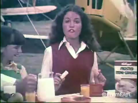 The still from the 1970s Kellogg's Shredded Wheat commercial features a lively outdoor breakfast scene. A young woman in a red vest, with light-colored sleeves, is animatedly speaking, likely promoting the cereal. Beside her, another girl is smiling and enjoying the moment. The table is set with a box of Frosted Mini-Wheats, milk, and juice, all suggesting a wholesome, family-friendly vibe. In the background, an airplane hints at a rural or adventurous setting, typical of the era's advertising aesthetics.