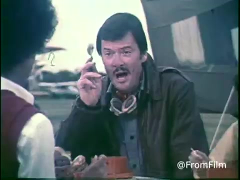 The still from the 'Kellogg's Shredded Wheat Commercial - 1970s' features a man with a mustache, dressed in a leather jacket and goggles, animatedly speaking while holding a spoon. He appears to be engaged in a conversation with others seated at a table, possibly outdoors. The setting includes an airplane in the background, suggesting a casual, lively atmosphere. The overall vibe captures the playful essence of 1970s commercials. Follow us on Twitter @FromFilm.