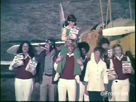 The still from the 1970s Kellogg's Shredded Wheat commercial features a group of diverse individuals gathered in front of an airplane. They wear matching outfits, predominantly in red and white, and are joyfully holding boxes of Shredded Wheat. A child is perched on an adult's shoulders, contributing to the playful and family-friendly vibe of the scene. The backdrop shows a rustic landscape, enhancing the nostalgic feel associated with this era's advertisements.
