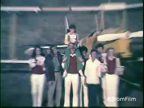 The still features a group of people posing together outdoors, with a yellow biplane visible in the background. Some participants are holding boxes of Kellogg's Shredded Wheat, showcasing a playful and family-friendly atmosphere. The attire reflects the casual style of the 1970s. This scene likely conveys a sense of joy and togetherness, typical of advertisements from that era.