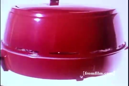 The still showcases a vintage red Mirro broiler, prominently displayed against a simple background. The design reflects the 1970s aesthetic, emphasizing both style and functionality. The rich color and polished surface convey a sense of retro charm typical of kitchen appliances from that era.