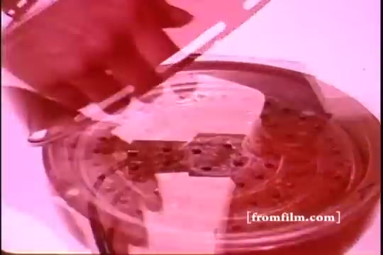 The still from the 'Mirro Broiler Commercial - 1970's' features a close-up of a hand preparing a dish, likely interacting with a broiler or cooking appliance. The colors are warm and slightly faded, typical of a 16mm print. The focus is on the broiler's grid or surface, suggesting a cooking demonstration. The overall aesthetic evokes a nostalgic, retro feel characteristic of 1970s advertisements.