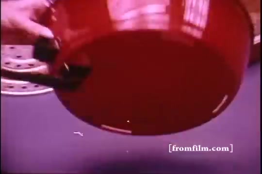 The still captures a close-up of a vibrant red cooking pot being held by a hand, showcasing its underside. The pot features a black handle and reflects a smooth surface typical of cookware from the 1970s. The background hints at a kitchen setting, with partially visible elements like a round rack, contributing to an overall retro aesthetic seen in vintage commercials.