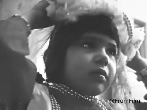The still from the 1968 American Cancer Society PSA features a young girl dressed in a decorative outfit. She appears to be adjusting a headpiece or veil while surrounded by a domestic setting. Her expression is thoughtful, possibly reflecting the significance of the moment. The black-and-white film captures the innocence and joy associated with childhood play, contrasting the serious health message of the PSA.