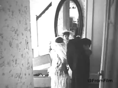 The still depicts a scene from a 1968 American Cancer Society PSA in black and white. It shows two individuals, likely in an intimate or domestic setting, with one person draped in a white garment or fabric. A large mirror in the background reflects parts of the environment, adding to the sense of depth. The overall atmosphere seems somber, in line with the serious message often conveyed in public service announcements about health issues like cancer.