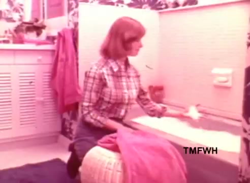 The still from the 'Rub-a-dub Dolly Commercial' features a woman sitting beside a bathtub, dressed in a plaid shirt and blue jeans. She appears to be preparing or cleaning the tub, surrounded by vibrant pink and patterned decor. The scene conveys a cheerful and inviting atmosphere typical of commercial aesthetics from that era. The visual style reflects the nostalgia of 16mm print quality.   For more vintage commercials, follow @FromFilm on Twitter!