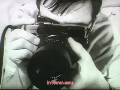 The still features a close-up of a man holding a camera, aiming it at the viewer. He appears focused and intent, capturing the essence of a moment. The image is likely in black and white, reflecting the style of the late 1960s. This shot contributes to the nostalgic and dynamic atmosphere often found in film trailers from that era.