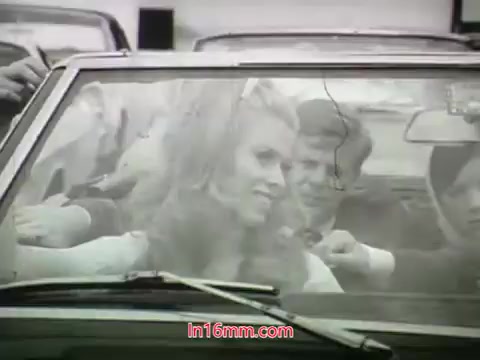 The still from the 1968 trailer for 'Out Of It' features a scene inside a convertible car. A woman with long, styled hair is smiling and seems to be engaged in conversation with a group of people outside the vehicle. The overall black-and-white aesthetic captures a vibrant moment, hinting at themes of youth and social interaction. The style reflects the era's cinematic trends.