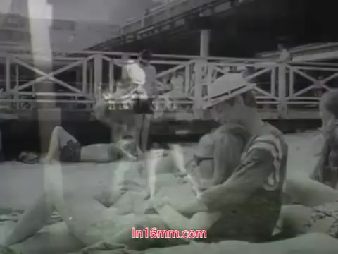 The still is a monochrome image from the 1968 trailer for 'Out Of It,' featuring a beach setting. People are lounging on the sand, with some engaged in casual activities, and a lively atmosphere is suggested. The focus is on youth and leisure, embodying the era's vibe. The presence of a pier and casual beach attire accentuates the film's exploration of relationships and experiences.