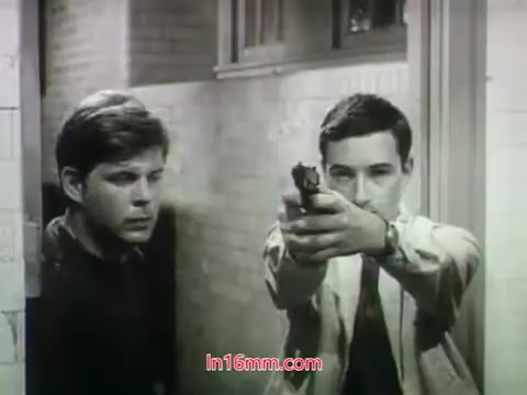 The still from the 1968 trailer for 'Out of It' features two young men in a tense moment. One character, looking concerned, stands slightly behind another who is pointing a handgun, suggesting a critical or dangerous situation. The stark black-and-white imagery enhances the drama of the scene.