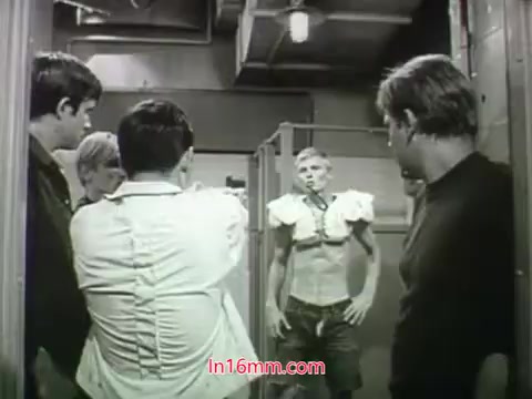 The still from the 1968 trailer of 'Out Of It' features a group of young men, some wearing casual clothing, staring at a character in the foreground. This character, standing in a somewhat awkward pose, is wearing a makeshift outfit that looks like padded shoulders and shorts. The scene conveys a mix of curiosity and amusement, encapsulating a comedic or absurd moment typical of the film's youthful and countercultural themes.
